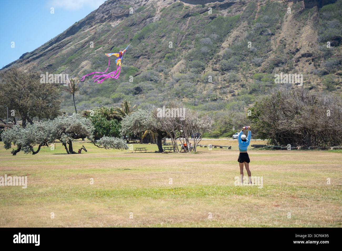 Eine Frau tanzt mit dem Wind, während sie einen Drachen allein an einem tropischen Strand fliegt, üppige grüne Berge, die sich hinter ihr in ruhiger Gelassenheit erheben. Stockfoto