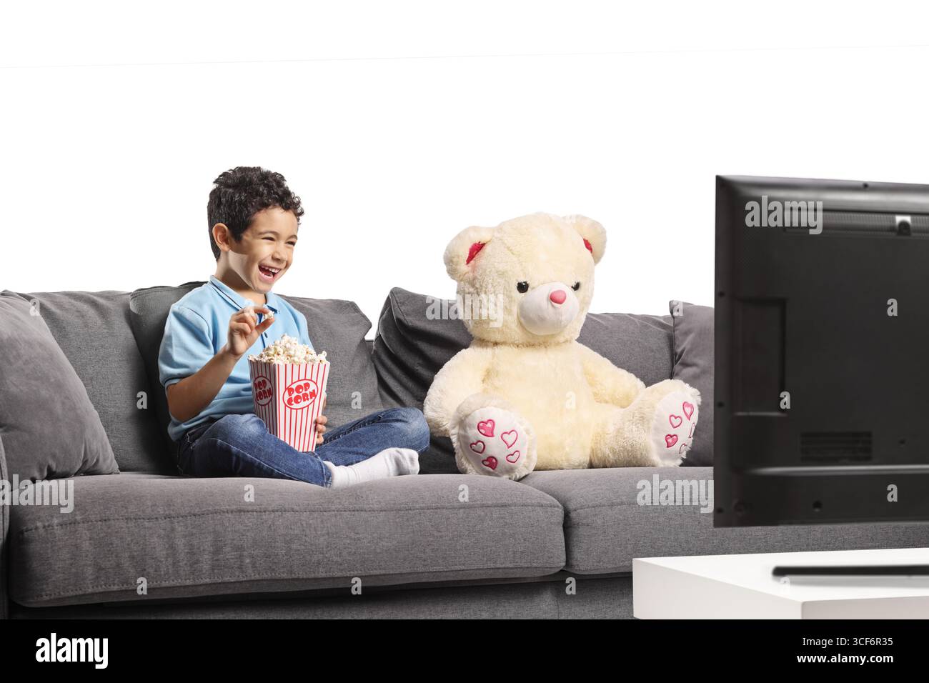 Glücklicher Junge, der Popcorn isst und mit seinem Teddybären fernsieht, isoliert auf weißem Hintergrund Stockfoto