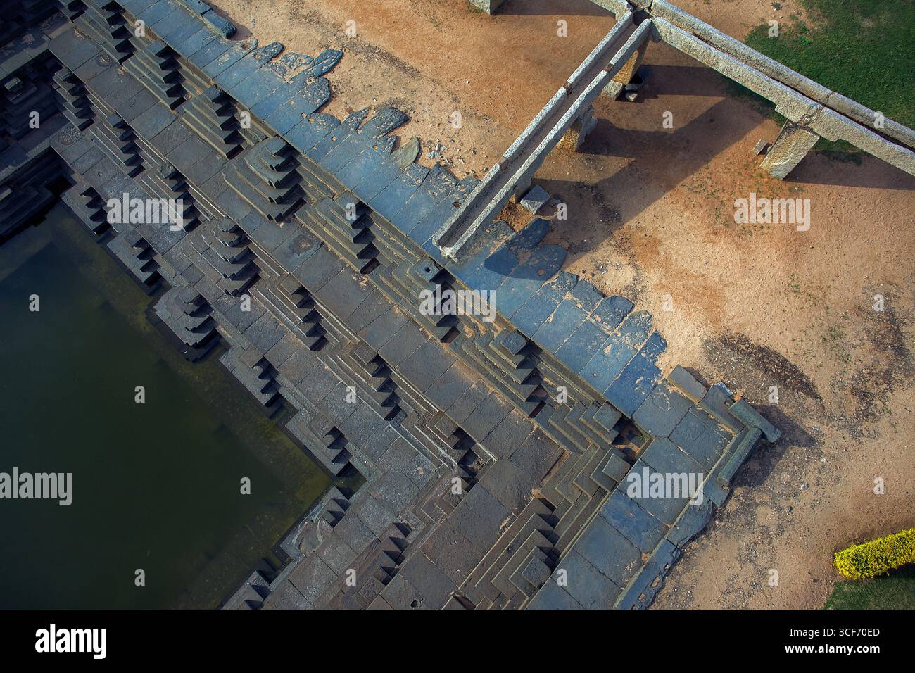 Die geometrische Präzision des abgestuften Tanks steht im Kontrast zu den organischen Texturen der umliegenden Landschaft und schafft eine faszinierende visuelle Harmonie, Hampi, Karnataka, Indien. Stockfoto