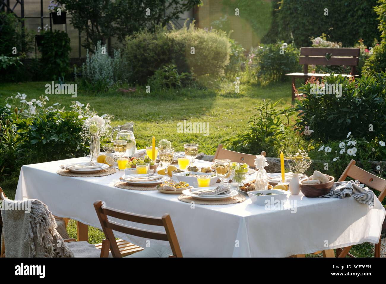 Gartentisch im Freien mit Tellern, Gläsern Orangensaft, Brot, Obst und Blumen auf Tischdecke, Holzstühlen um den Tisch herum, üppiges Grün im Hintergrund Stockfoto