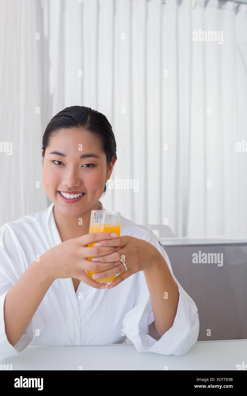 Koreanische Erwachsene Frau, die am Esstisch sitzt und ein Glas Orangensaft vor dem Hintergrund des Vorhangs hält Stockfoto
