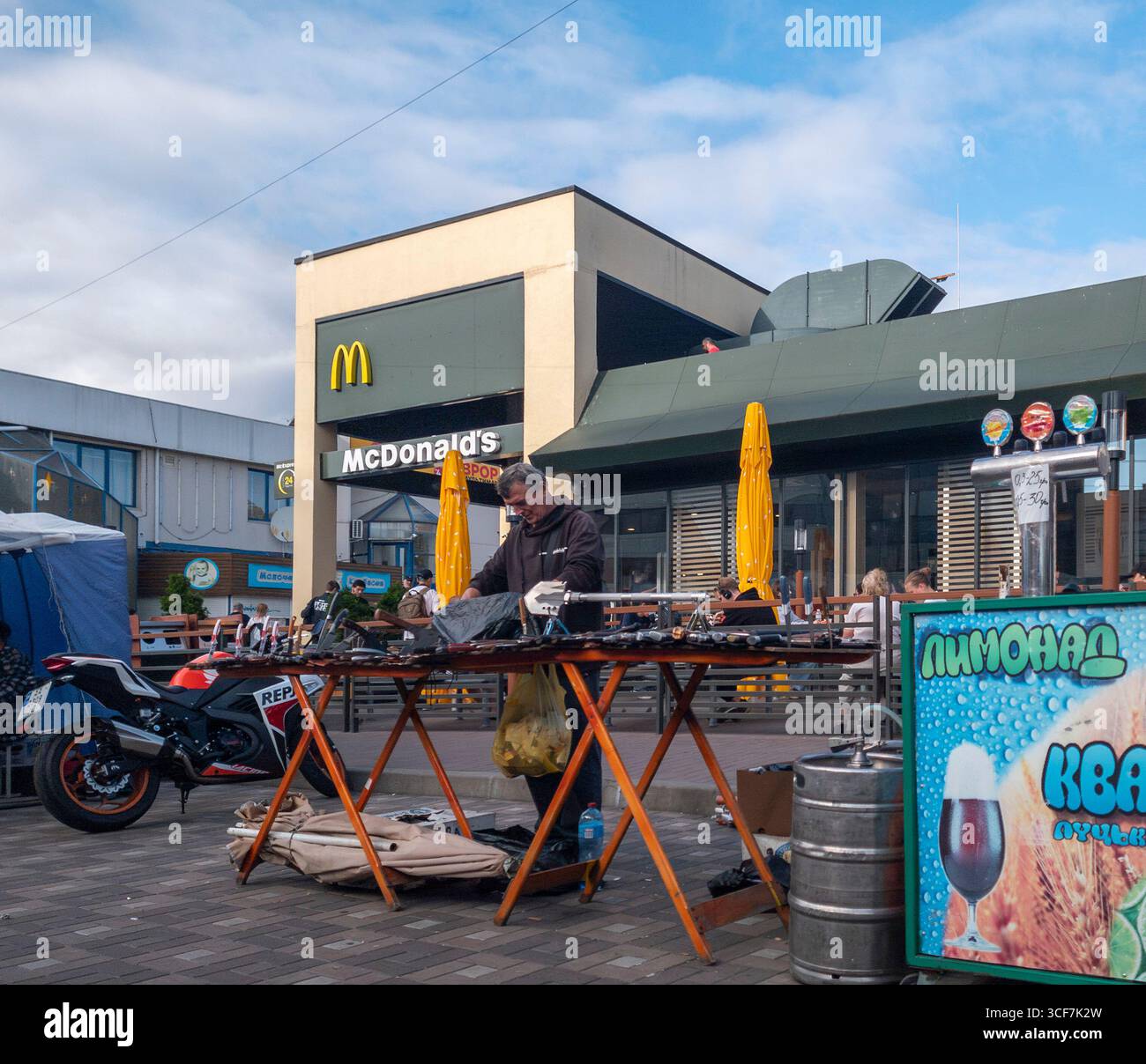 Kiew, Ukraine - 20. Juli 2025: Straßenverkäufer arrangiert seine Waren an einem Stand vor McDonald's, ein Motorrad parkt in der Nähe und die Leute genießen ihre Mahlzeiten auf der Terrasse Stockfoto