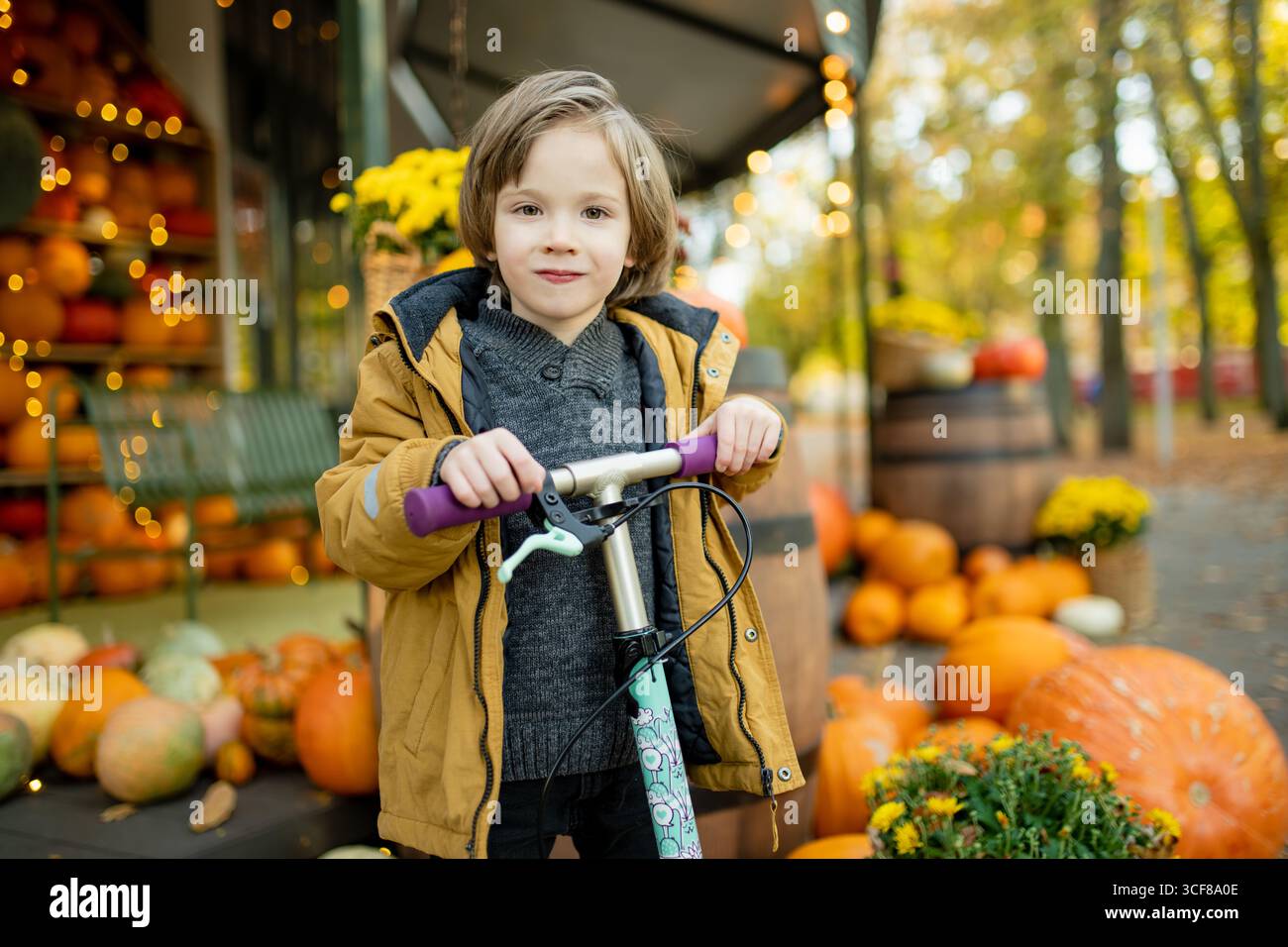 Ein fröhliches Kind steht mit einem Roller neben einem festlichen Herbstkürbis, umgeben von bunten Kürbissen und goldenen Blättern Stockfoto