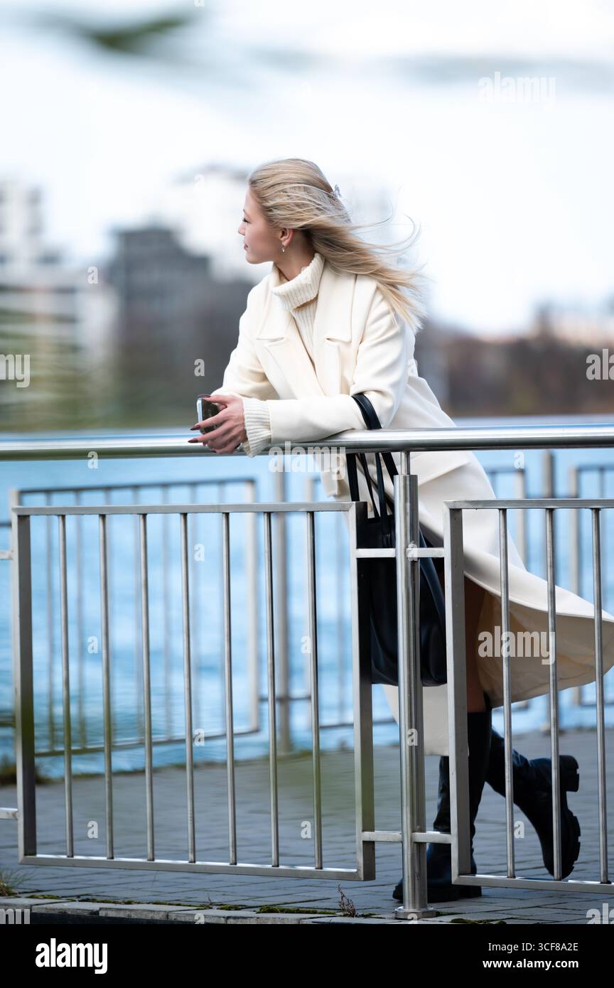 Eine stilvolle Frau lehnt sich an ein Geländer, ihr Haar tanzt im Wind, während sie den Blick auf das Wasser in einer geschäftigen Stadt bewundert. Die Szene vermittelt Ruhe Stockfoto