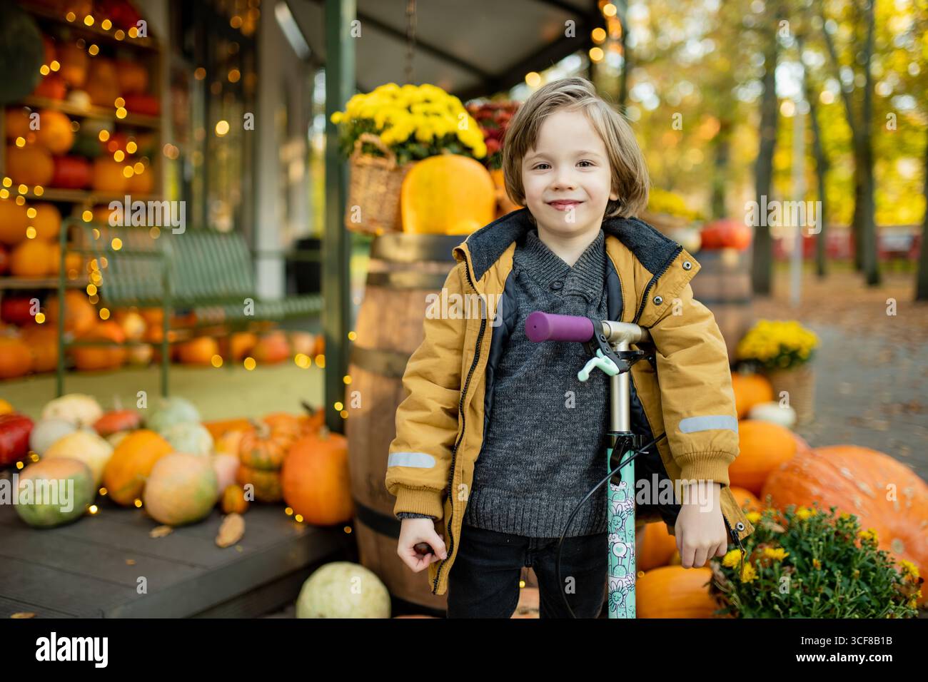 Ein fröhliches Kind steht mit einem Roller neben einem festlichen Herbstkürbis, umgeben von bunten Kürbissen und goldenen Blättern Stockfoto