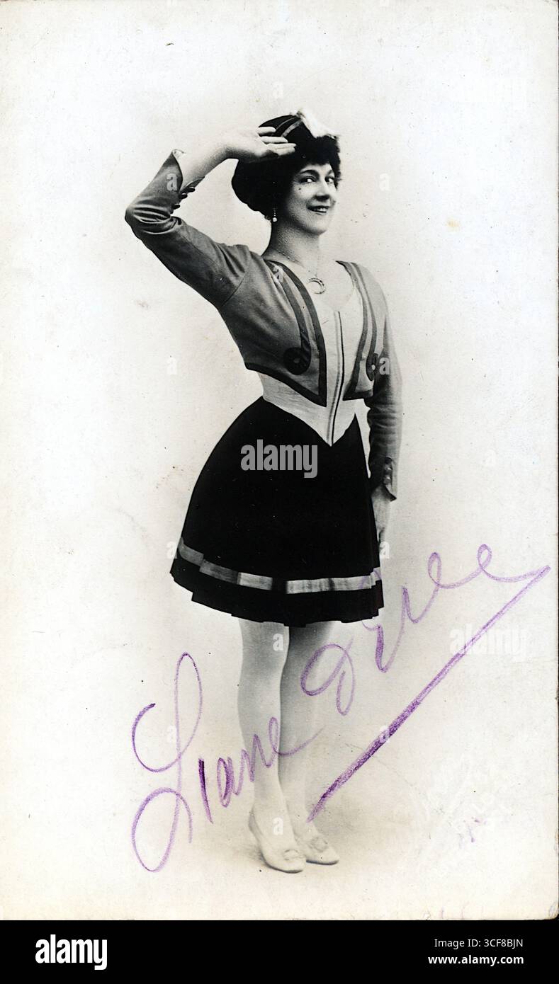 Eine poetische Figur steht in einem theatralischen Gruß, in einem maßgeschneiderten Ensemble aus Jacke und Rock, das an edwardisches Bühnenkostüm erinnert. Weiße Strumpfhosen und Tanzschuhe erinnern an Ballett- oder Kabarett-Einflüsse, während die dekorativen Bänder am Saum rhythmisches Flair verleihen. Die Signatur „Liane Dire“ bewegt sich in violetter Tinte über den unteren Rand und verankert das Bild in einer persönlichen und performativen Identität. Wahrscheinlich aus den 1910er Jahren, trägt das Foto die charakteristischen Merkmale der Bold Street, Liverpool Studio Porträts – gestochen scharf, ausdrucksstark und kulturell resonant. Stockfoto