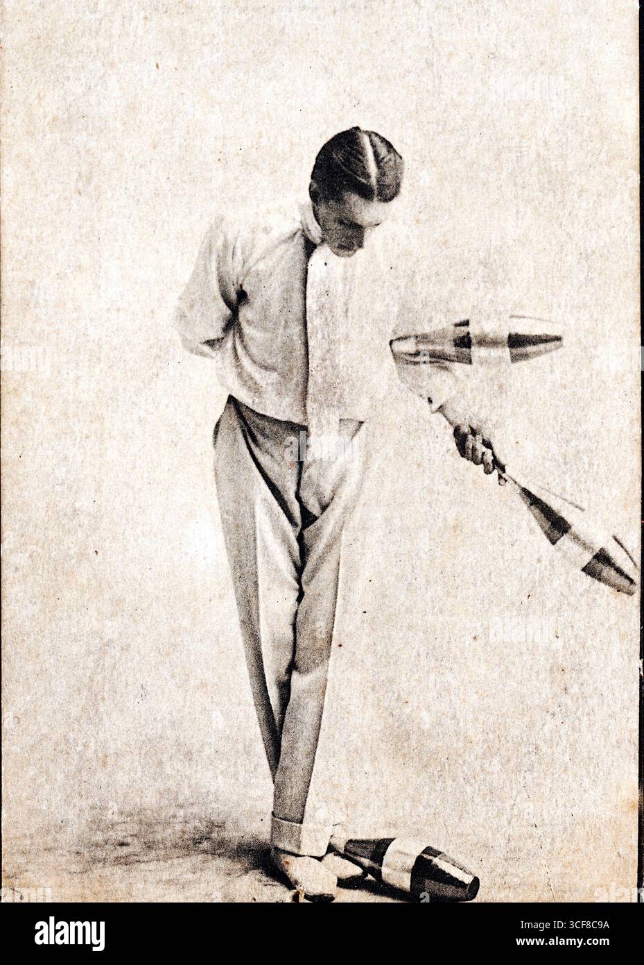 Leolin, ein hochkarätiger Edwardianer Jongleur, steht in formeller Kleidung mitten im Trend: Weißes Hemd, Fliege und Hosen mit hohem Bund. Er hält zwei Schläger in der rechten Hand, während ein dritter in der Nähe seiner Füße liegt, was auf einen Moment der Pause oder des Übergangs hindeutet. Sein linker Arm ist hinter seinem Rücken verstaut, und sein Blick nach unten zeigt konzentrierte Konzentration. Der schlichte, strukturierte Hintergrund unterstreicht seine einsame Präsenz. Diese Postkarte wurde von Imperial Publishing Co. Of Longton, Staffels, 1910er veröffentlicht und fängt die Eleganz und Disziplin der frühen Unterhaltungswelt ein. Stockfoto