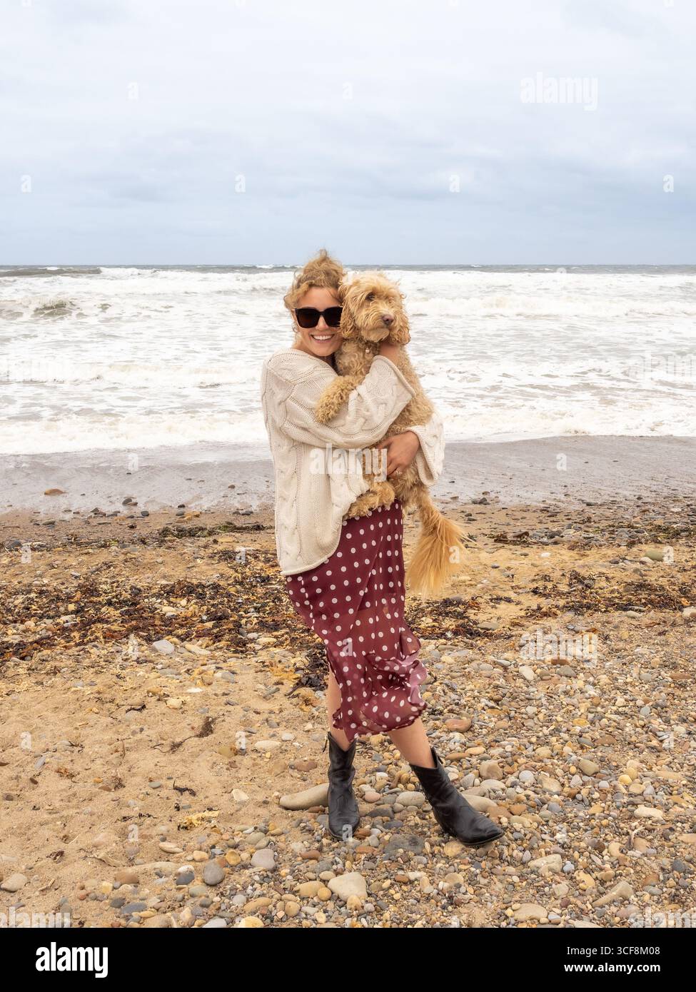 Eine stilvoll gekleidete Frau hält ihren Hund Cockapoo, während sie am Sand- und Kiesstrand von Saltburn-by-the-Sea, North Yorkshire, Großbritannien, steht. Stockfoto