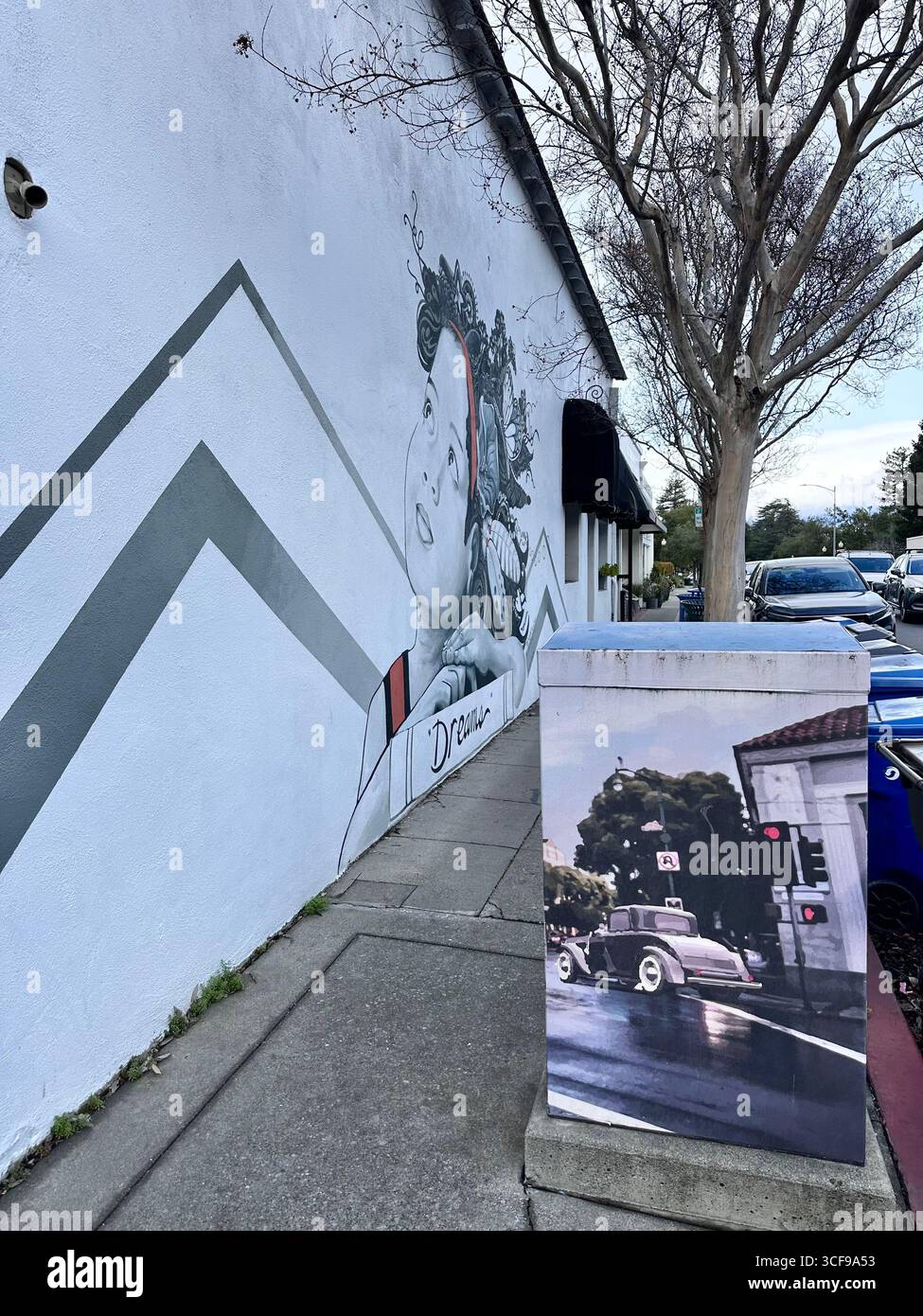 Ein Straßenbild, das ein Kind mit aufwendigen Haarmotiven zeigt, neben einer bemalten Gebrauchskiste, die einen Oldtimer und eine historische Bauszene zeigt - Smartphone-aufgenommenes Stockfoto