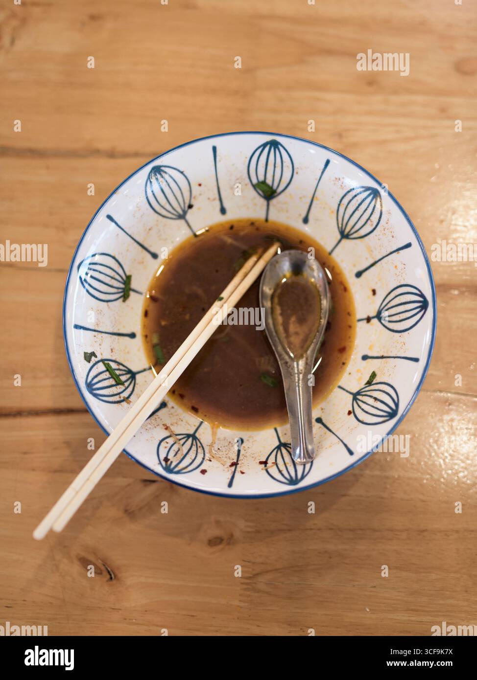 Blick von oben auf eine leere Nudelsuppe mit übrig gebliebener Brühe, Essstäbchen und Metalllöffel auf einem Holztisch. Minimalistisches Speisekonzept, asiatische Straße Stockfoto