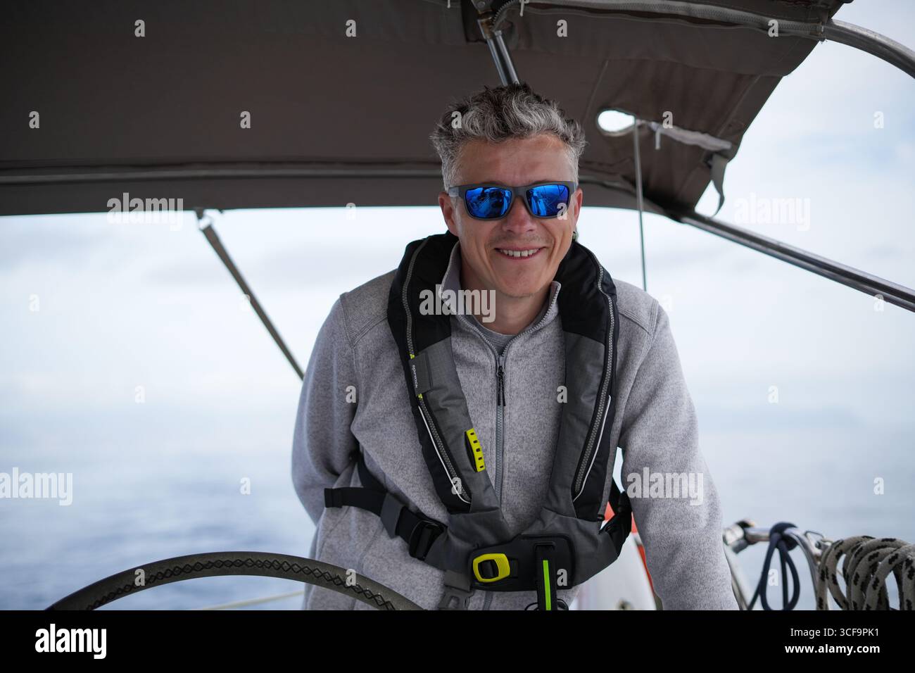 Porträt eines Mannes, der auf einer Yacht mit Schwimmweste und Sonnenbrille segelt und am Ruder mit Meer und Horizont in der Nähe der Kanarischen Inseln Spanien lächelt Stockfoto