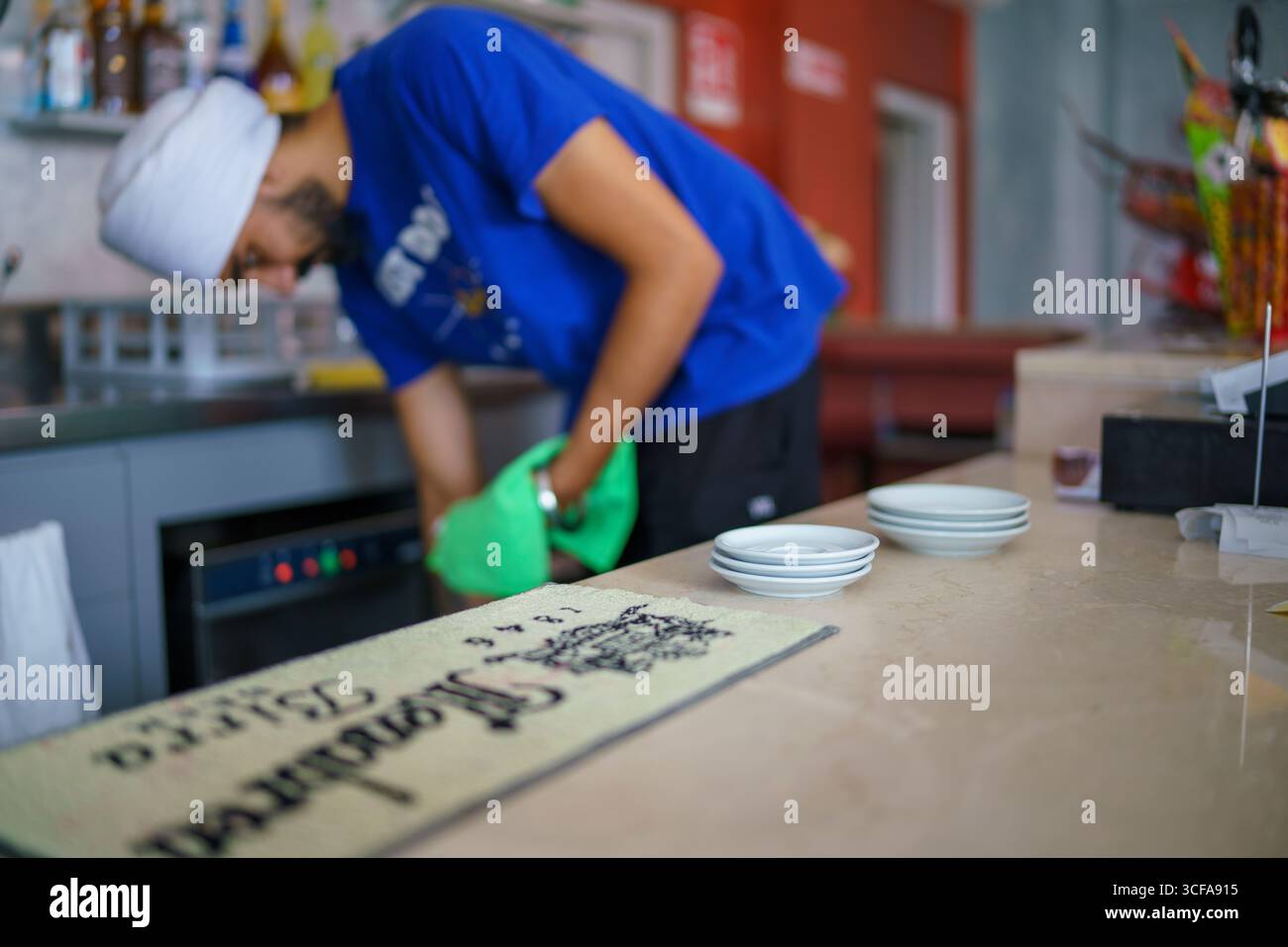 Cremona, Italien 25. Juli 2025 der Bartender wischt die Bar mit einem grünen Tuch ab, mit einem Stapel Untertassen und einem Estrella Damm Tischset Stockfoto