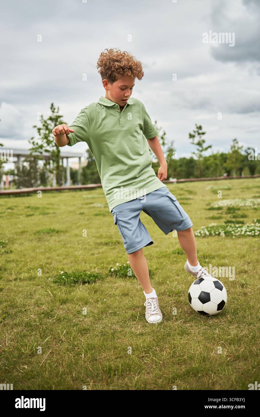 Kaukasischer Junge, der im Freien Fußball spielt, Ball auf einem grasbewachsenen Feld mit Bäumen im Hintergrund tritt, aktive Bewegung und Konzentration zeigt, in legeren Kleidern Stockfoto