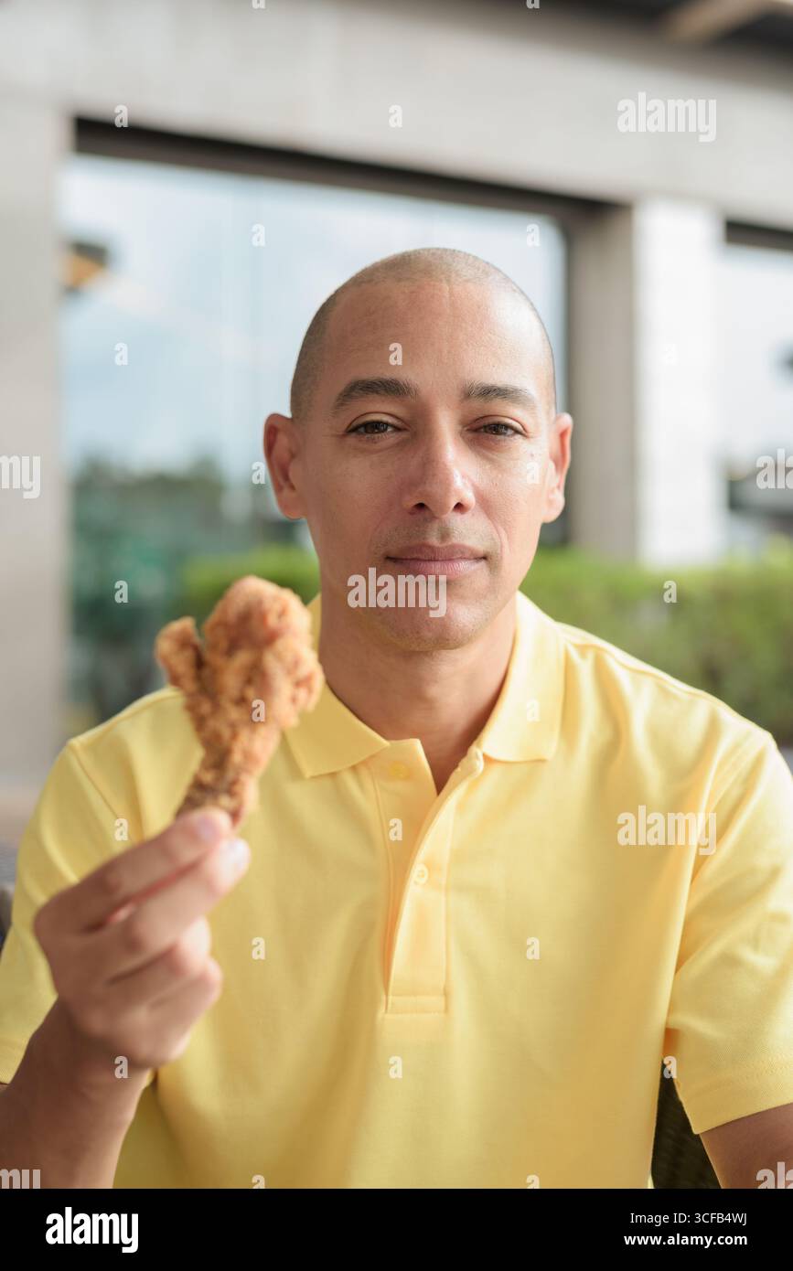 Porträt eines hispanischen Mannes mittleren Alters, der draußen sitzt und während des Mittagessens knuspriges gebratenes Huhn isst, während er einen entspannten Lebensstil in einer ungezwungenen Outdoor-Umgebung genießt Stockfoto