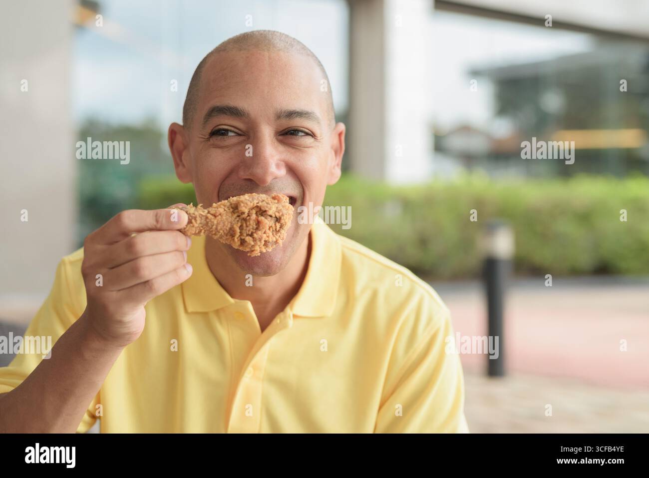 Porträt eines hispanischen Mannes mittleren Alters, der draußen sitzt und während des Mittagessens knuspriges gebratenes Huhn isst, während er einen entspannten Lebensstil in einer ungezwungenen Outdoor-Umgebung genießt Stockfoto