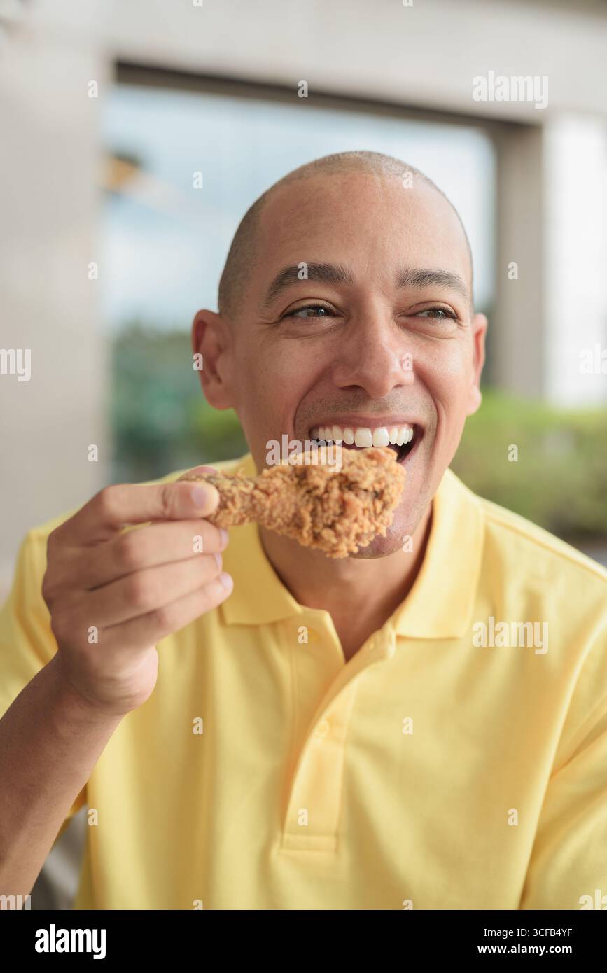 Porträt eines hispanischen Mannes mittleren Alters, der draußen sitzt und während des Mittagessens knuspriges gebratenes Huhn isst, während er einen entspannten Lebensstil in einer ungezwungenen Outdoor-Umgebung genießt Stockfoto