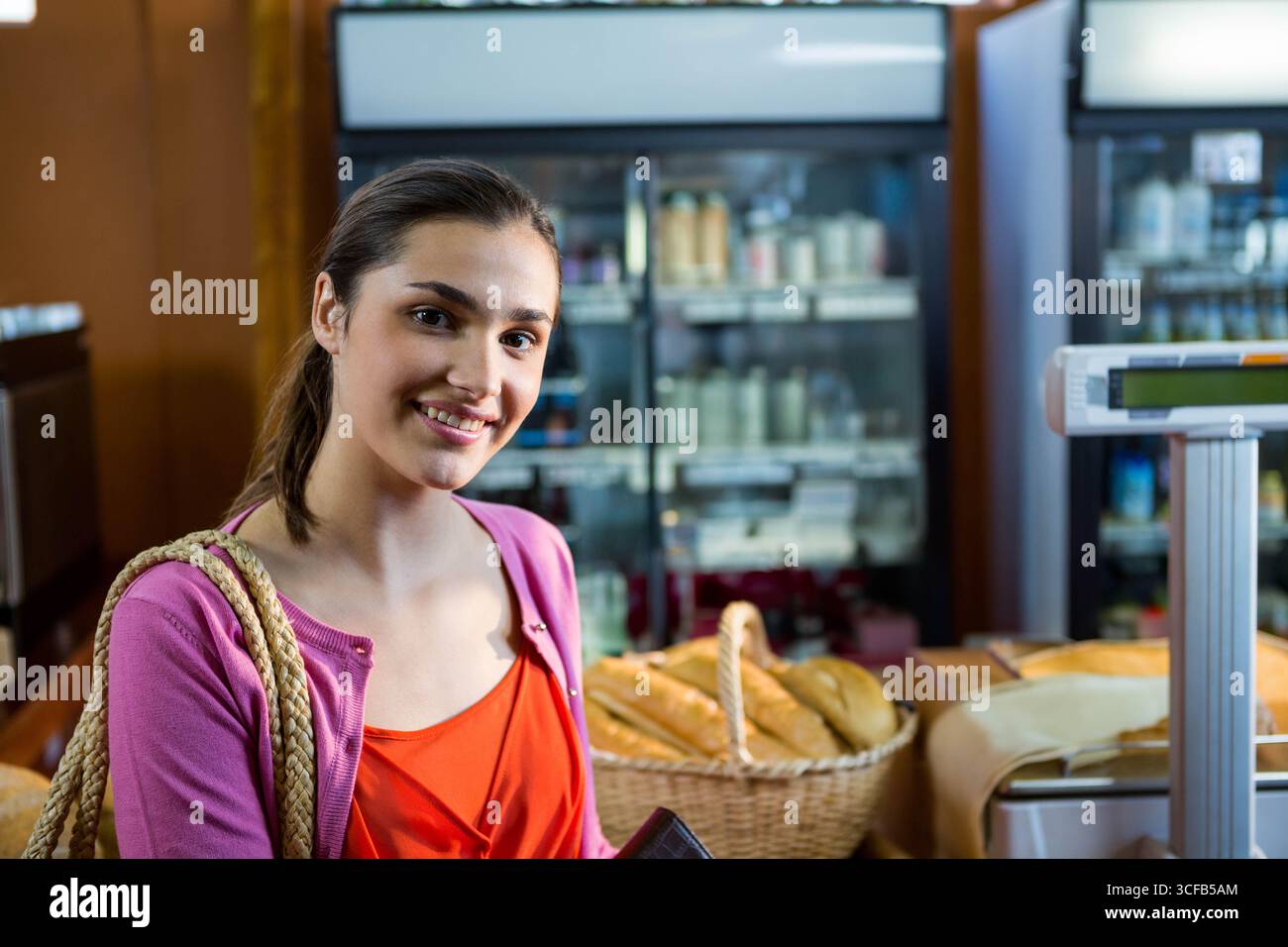 Weibliche Käuferin steht in der Bäckerei mit Brieftasche, Einkaufstasche neben Brotkörben, digitale Waage Stockfoto