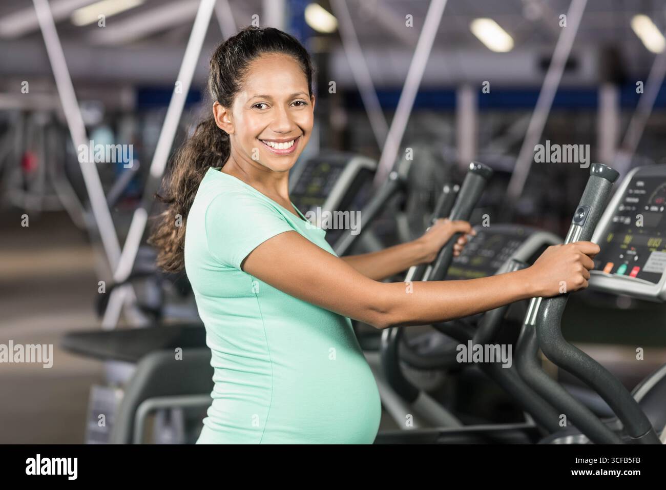 Schwangere Frau, die im Fitnessstudio mit Konsole und Kardiogeräten auf einem Crosstrainer steht Stockfoto