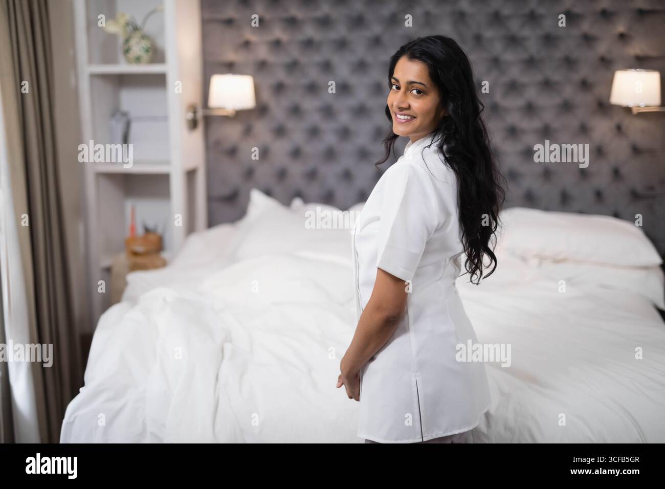Indische Frau, die neben dem Bett steht, mit weißer Bettwäsche, getuftetem Kopfteil und Lampen im Hotel, Kopierraum Stockfoto