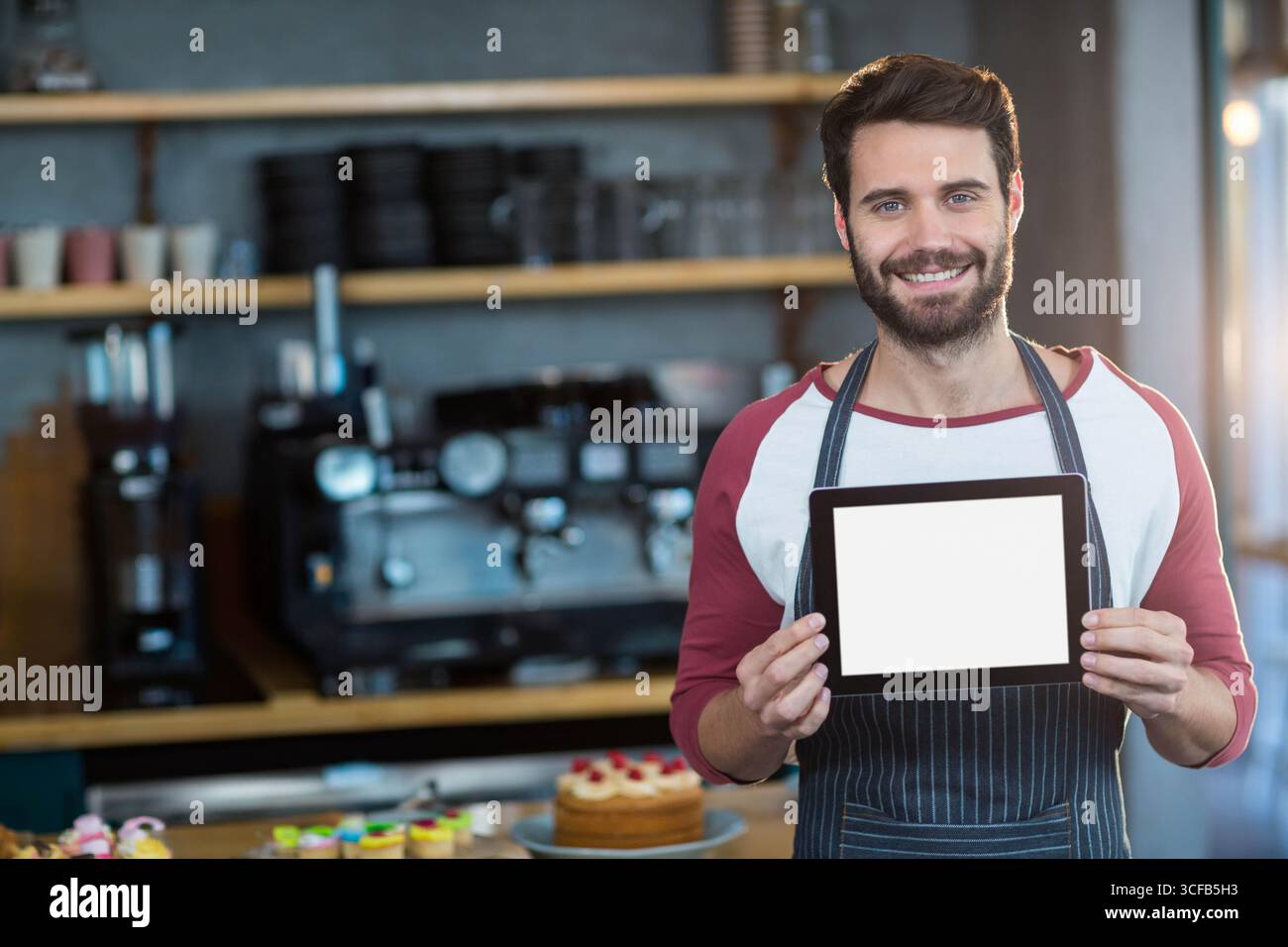 Bärtiger Mann Barista, der ein Tablet hält, das im Café mit Gebäck und Espressomaschine steht Stockfoto