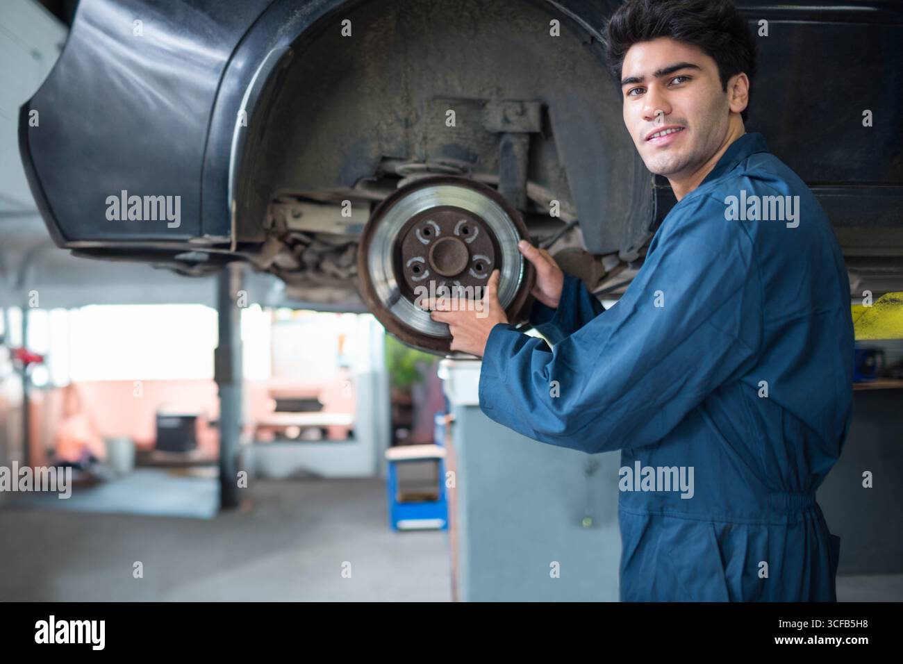 Asiatischer Mann, der Overalls trägt und den Bremsrotor auf hydraulischem Aufzug in der Reparaturwerkstatt überprüft, Kopierraum Stockfoto