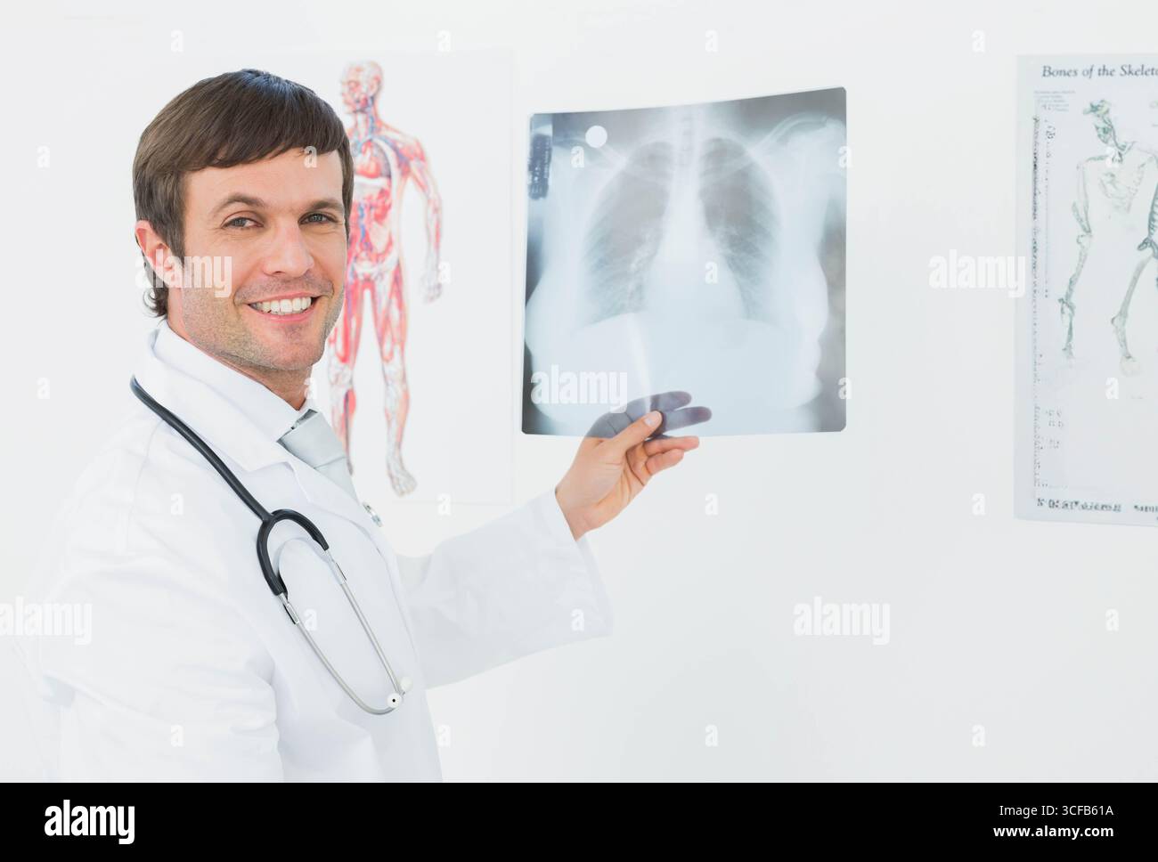 Männlicher Arzt mit Thoraxröntgenfilm auf Augenhöhe im Untersuchungsraum mit Stethoskop und Plakaten Stockfoto