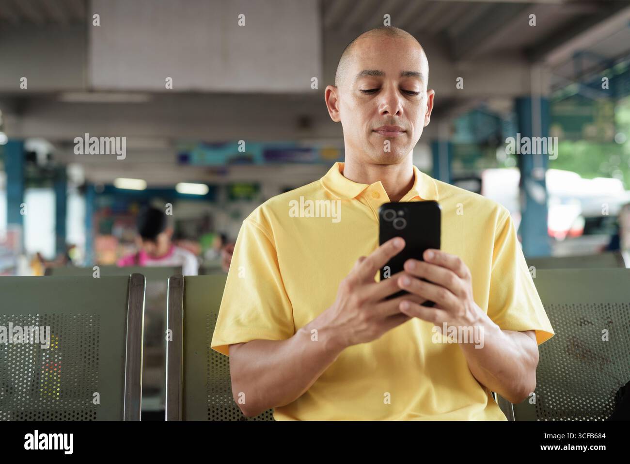 Reifer kahlköpfiger hispanischer Mann, der an einem Busbahnsteig im Freien sitzt und auf den Transport wartet, während er sein Smartphone durchsucht. Reisen, Pendeln und Stockfoto