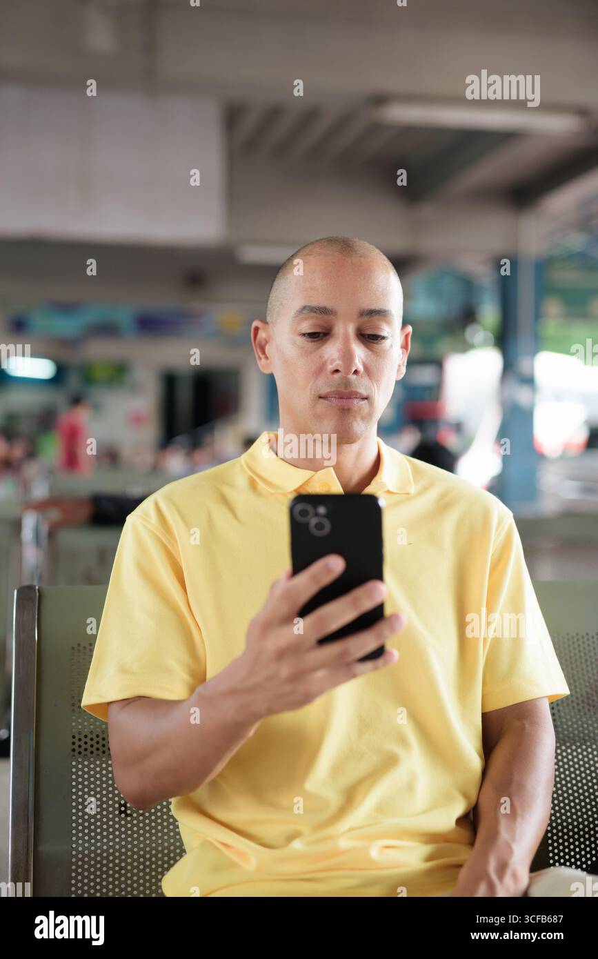 Reifer kahlköpfiger hispanischer Mann, der an einem Busbahnsteig im Freien sitzt und auf den Transport wartet, während er sein Smartphone durchsucht. Reisen, Pendeln und Stockfoto