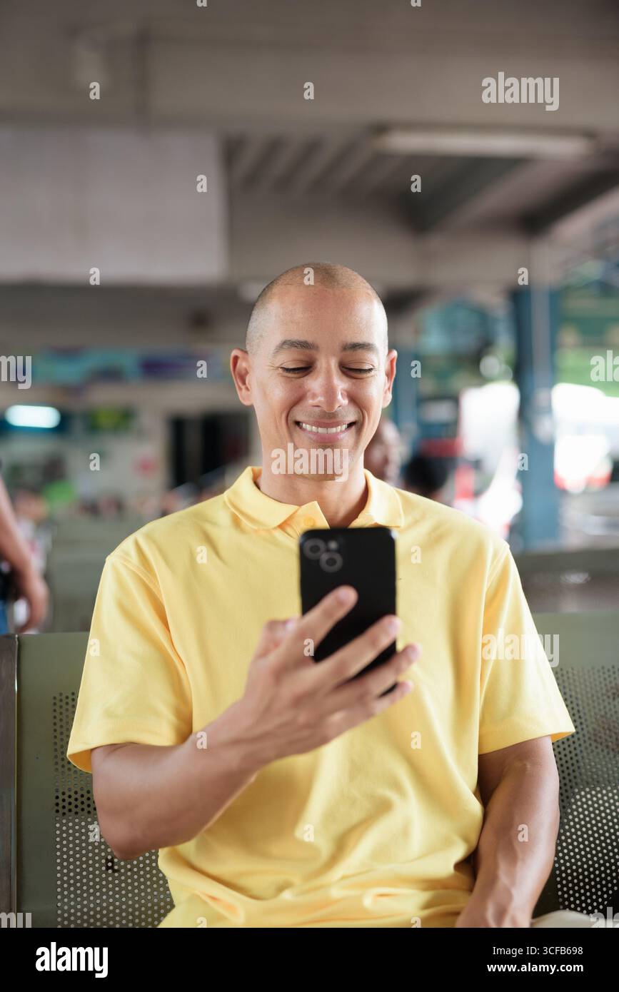 Reifer kahlköpfiger hispanischer Mann, der an einem Busbahnsteig im Freien sitzt und auf den Transport wartet, während er sein Smartphone durchsucht. Reisen, Pendeln und Stockfoto