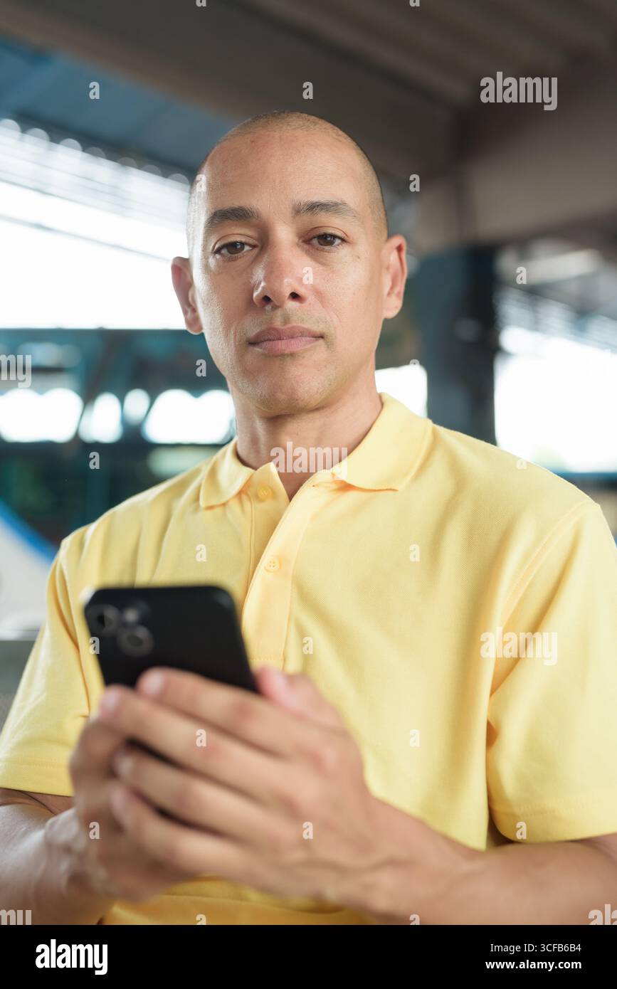 Reifer kahlköpfiger hispanischer Mann, der an einem Busbahnsteig im Freien sitzt und auf den Transport wartet, während er sein Smartphone durchsucht. Reisen, Pendeln und Stockfoto