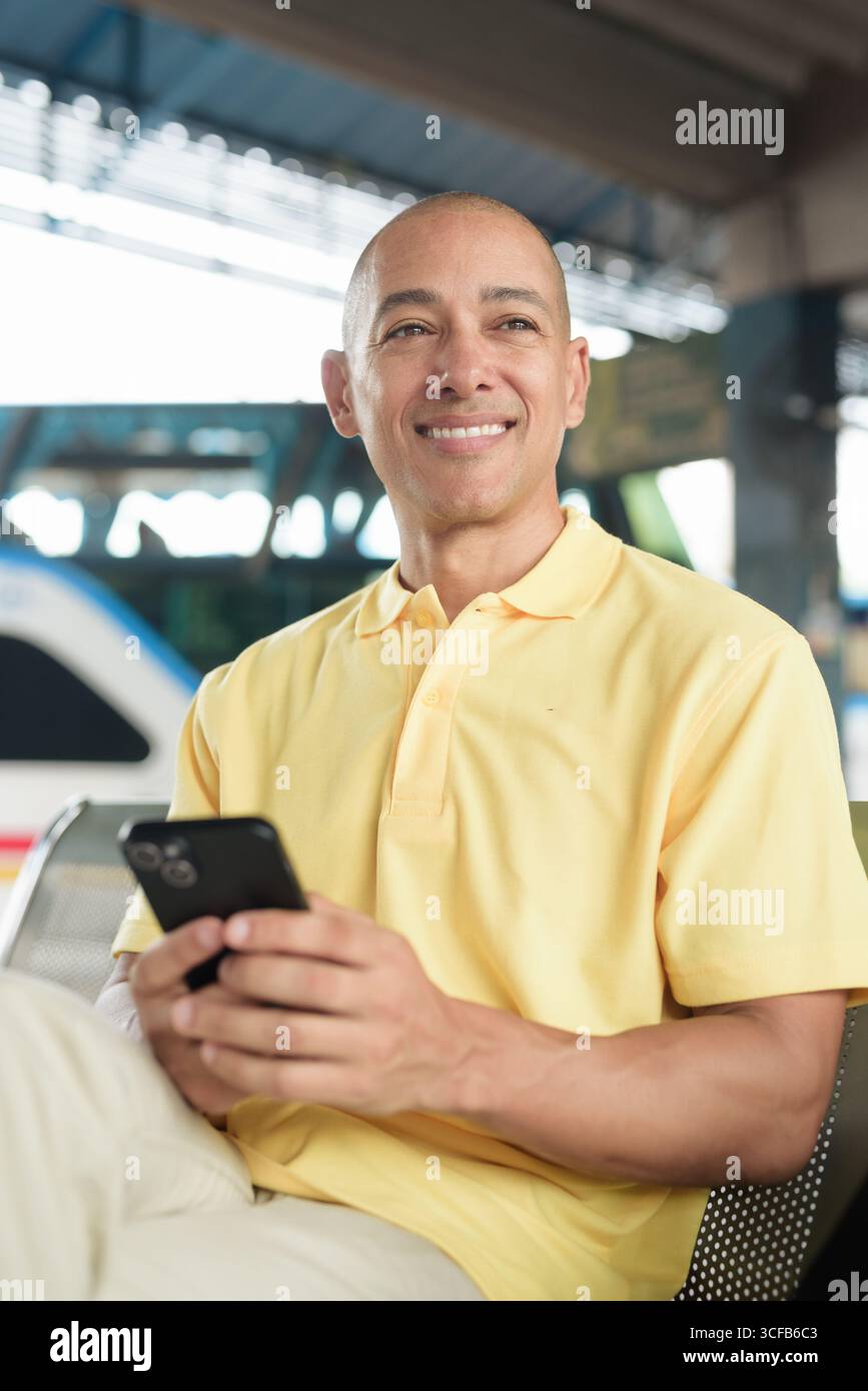 Reifer kahlköpfiger hispanischer Mann, der an einem Busbahnsteig im Freien sitzt und auf den Transport wartet, während er sein Smartphone durchsucht. Reisen, Pendeln und Stockfoto