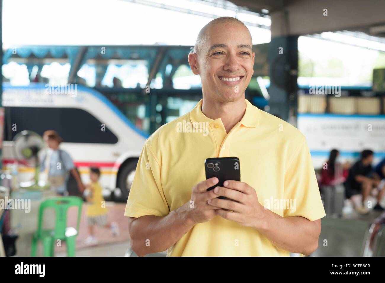 Reifer kahlköpfiger hispanischer Mann, der an einem Busbahnsteig im Freien sitzt und auf den Transport wartet, während er sein Smartphone durchsucht. Reisen, Pendeln und Stockfoto