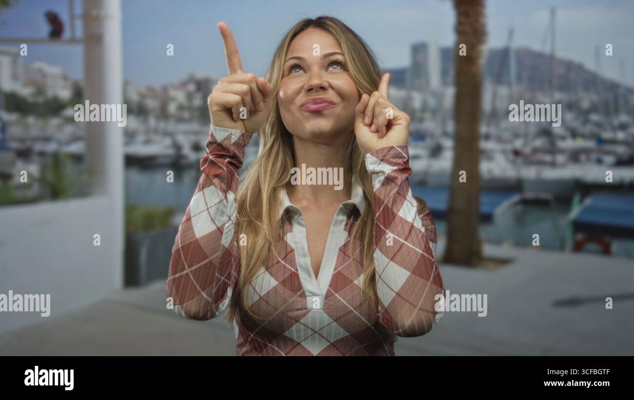 Eine blonde Frau mit einem fröhlichen Lächeln zeigt beide Zeigefinger nach oben auf die Marina Dock; Erwartungsoptimist. Stockfoto