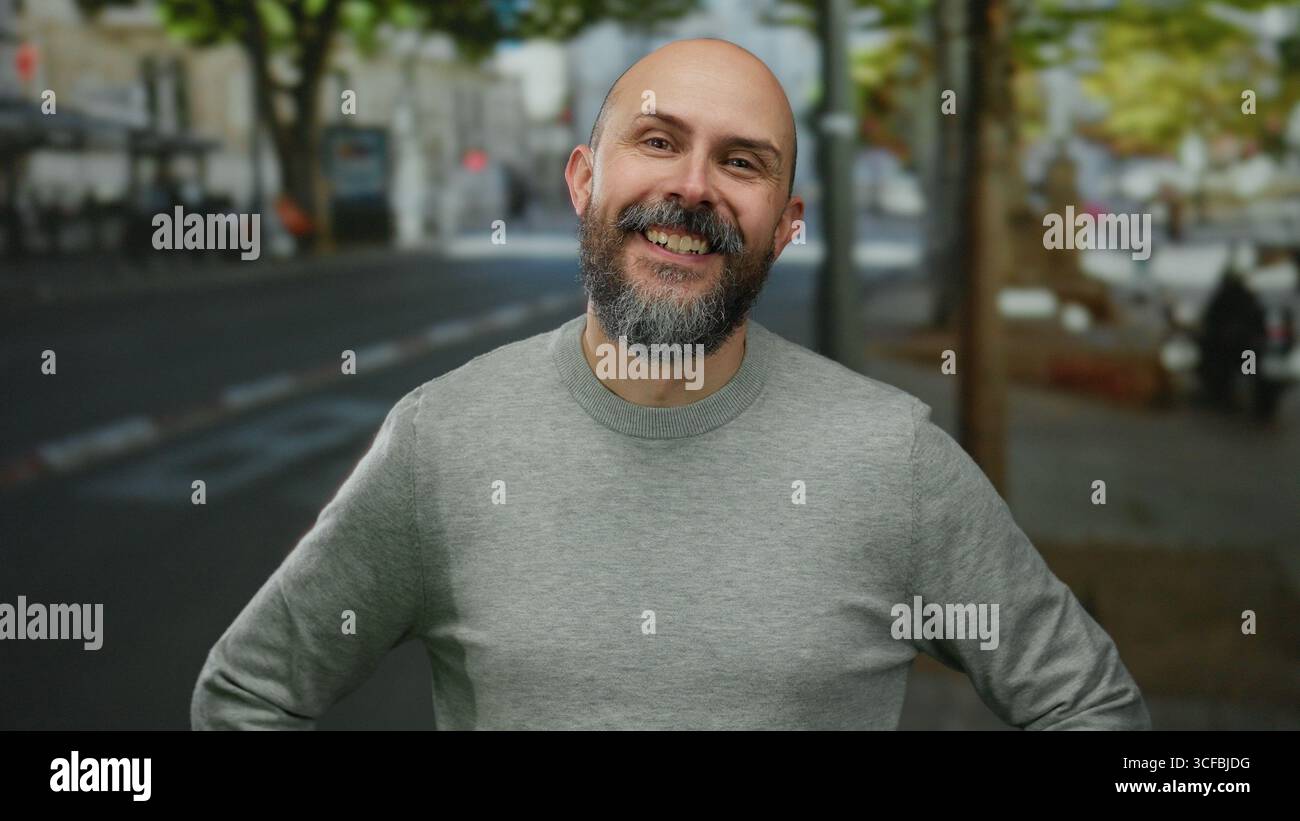 Bärtiger Mann in urbaner Umgebung mit grauem Pullover, der tagsüber auf einer von Bäumen gesäumten Stadtstraße warm lächelt und eine freundliche Outdoor-Atmosphäre darstellt. Stockfoto
