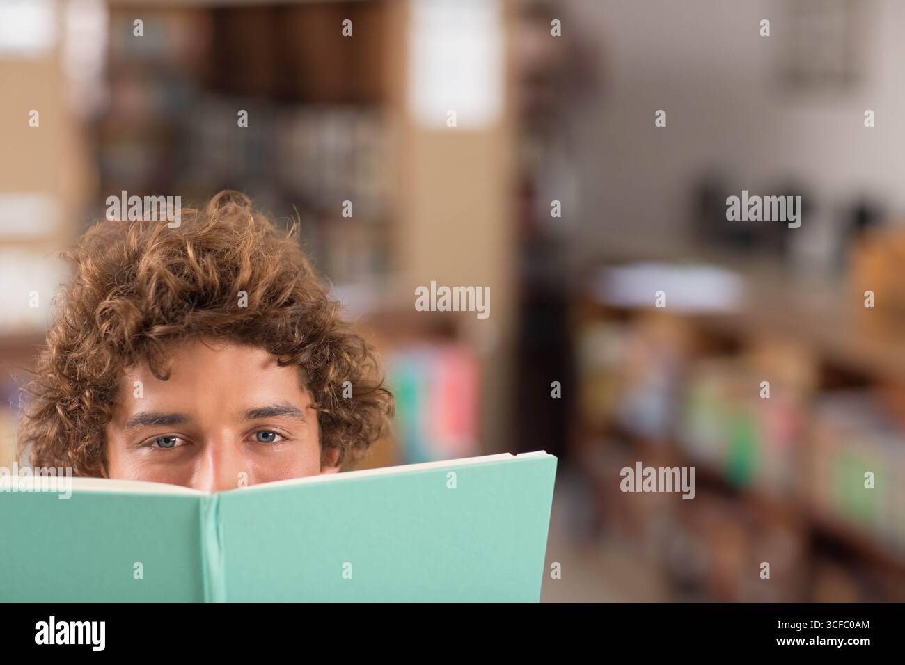 Mann, der über hellgrünes Hardcover-Buch in der Bibliothek mit Holzregalen, Tischen und Stühlen blickt Stockfoto