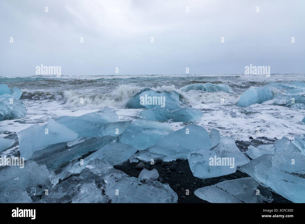 Blick auf die kristallklaren Eisfelsen, die entlang des schwarzen Sandstrandes verstreut sind, wo die schaumigen Wellen unter dem bewölkten Himmel krachen, Vik, Island. Stockfoto