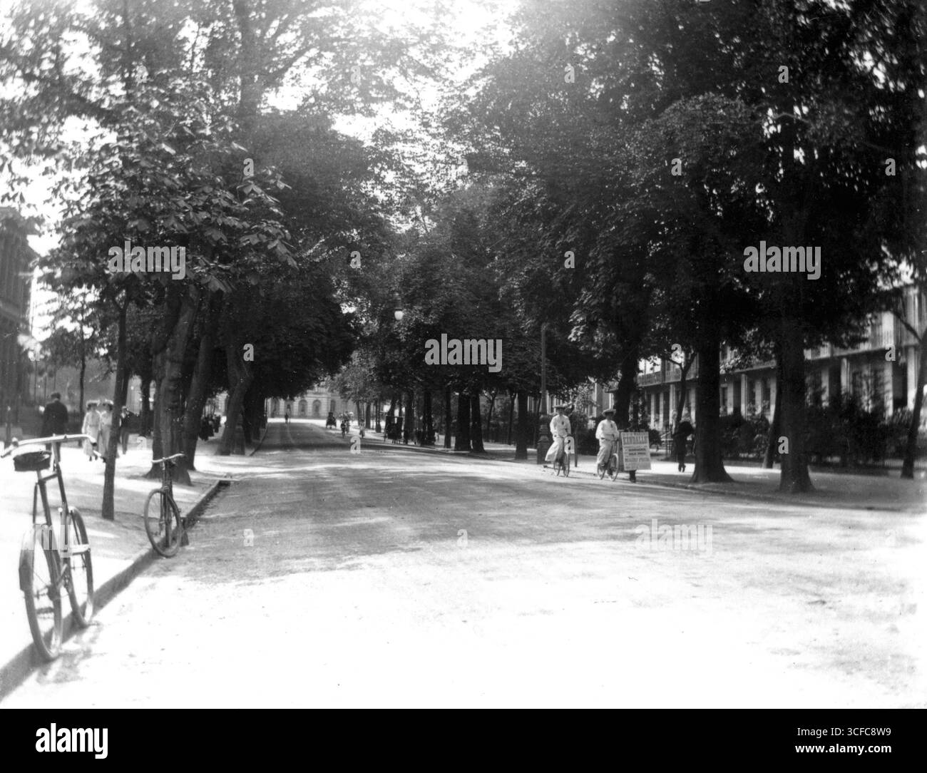 Gloucestershire, England. Um 1905 – ein Edwardianfoto der Promenade, Cheltenham. Fußgänger laufen auf den Gehwegen und Radfahrer benutzen die Straße. Ein Mann läuft die Straße entlang und trägt ein Sandwichbrett, das für den Verkauf von Sonnenschirmen bei Kendalls wirbt. Dieses Unternehmen befindet sich in der Cheltenham High Street und ist auf Sonnenschirme, Gehstöcke und Sonnenschirme spezialisiert. Am Ende der Promenade ist das Queen's Hotel zu sehen. Stockfoto