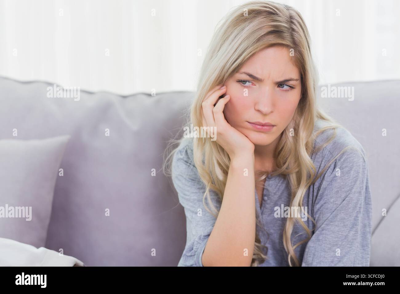 Frau, die zu Hause auf dem Sofa sitzt, Kopf auf der Hand mit Kissen in der Nähe von Vorhängen, Kopierraum Stockfoto
