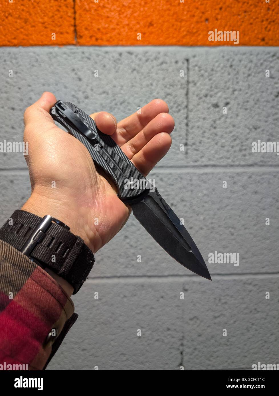 Die Hand eines Mannes in einem lässigen karierten Hemd hält ein offenes schwarzes, taktisches Klappmesser fest, ein unverzichtbares Alltagsgegenstand. Stockfoto