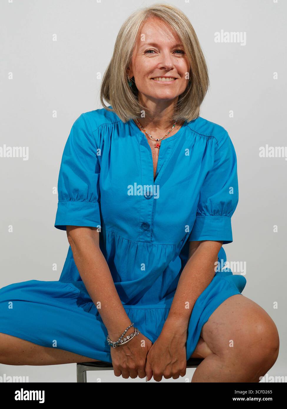 Lächelnde Frau in einem hellblauen Kleid, die auf einem Hocker vor einem schlichten Hintergrund sitzt. Italien Stockfoto