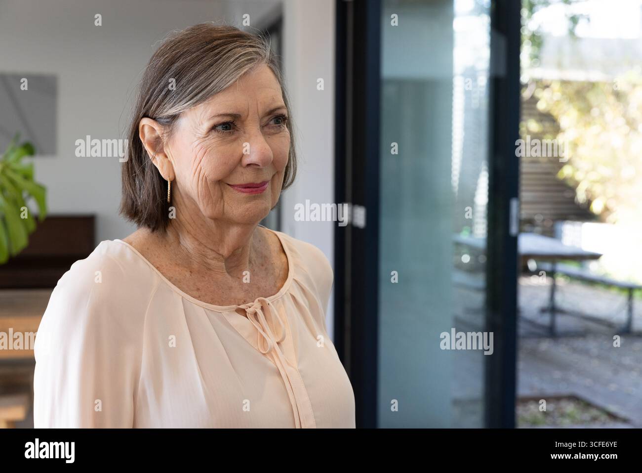 Seniorin steht im Esszimmer mit Holztisch in der Nähe der Glasschiebetür, Kopierraum Stockfoto