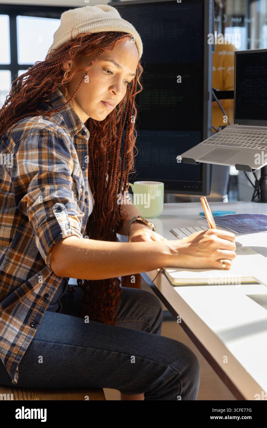Eine afroamerikanische Softwareentwicklerin sitzt am Schreibtisch und macht Notizen neben zwei Monitoren Stockfoto