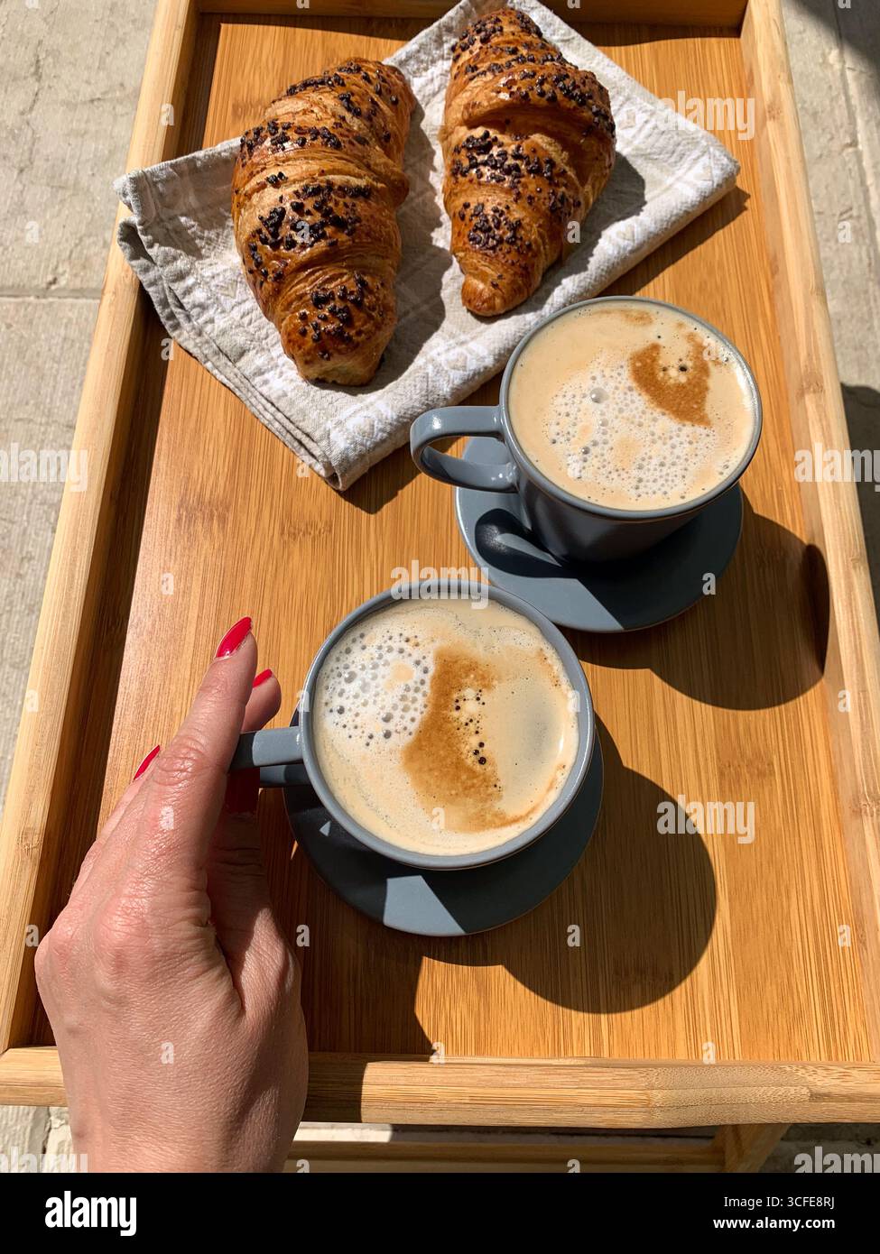 Traditionelles italienisches Frühstück. Frische Croissants und Cappuccino auf einem Holztablett. Frau Hand hält eine Tasse Cappuccino, Blick von oben. - Smartphone-aufgenommenes Stockfoto