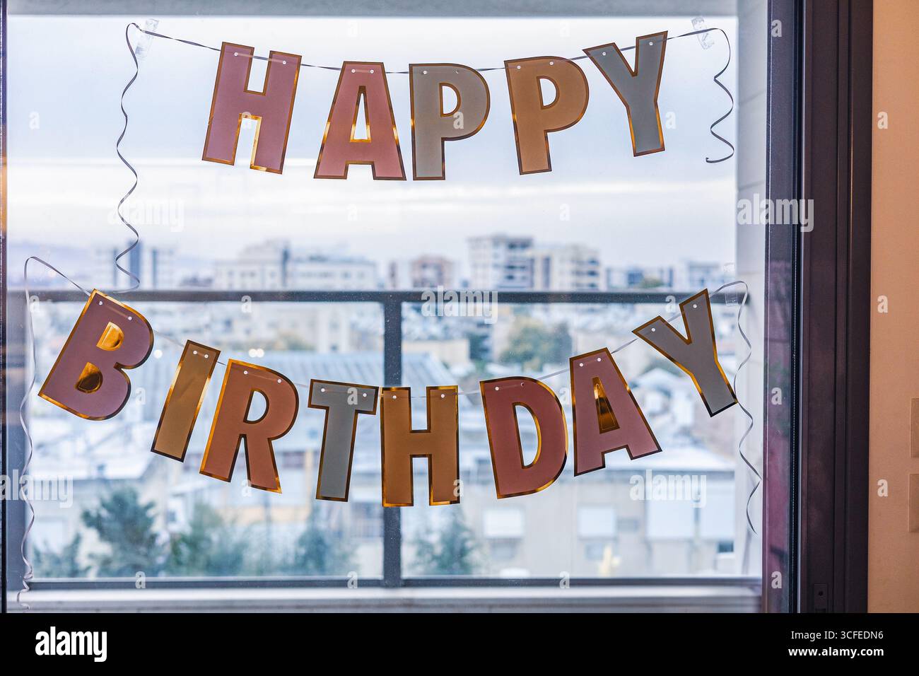 Nahaufnahme des Happy Birthday-Banners, das an Fensterglas hängt, mit verschwommenem Stadtbild im Hintergrund Stockfoto