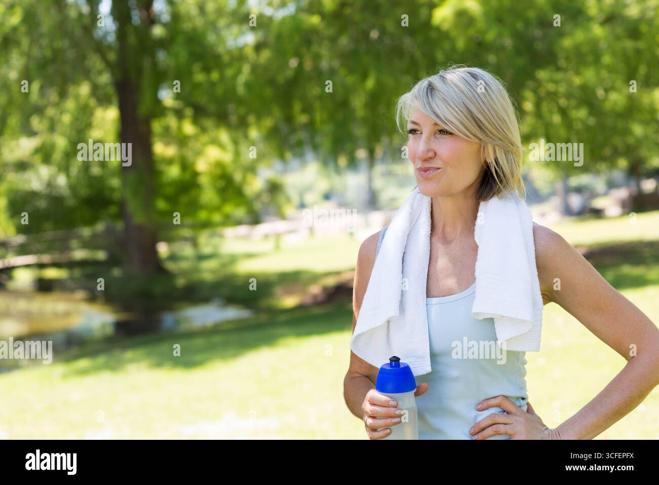 Eine ältere Frau, die im sonnigen Park neben dem Teich steht und eine Wasserflasche mit Tanktop und Kopierraum hält Stockfoto