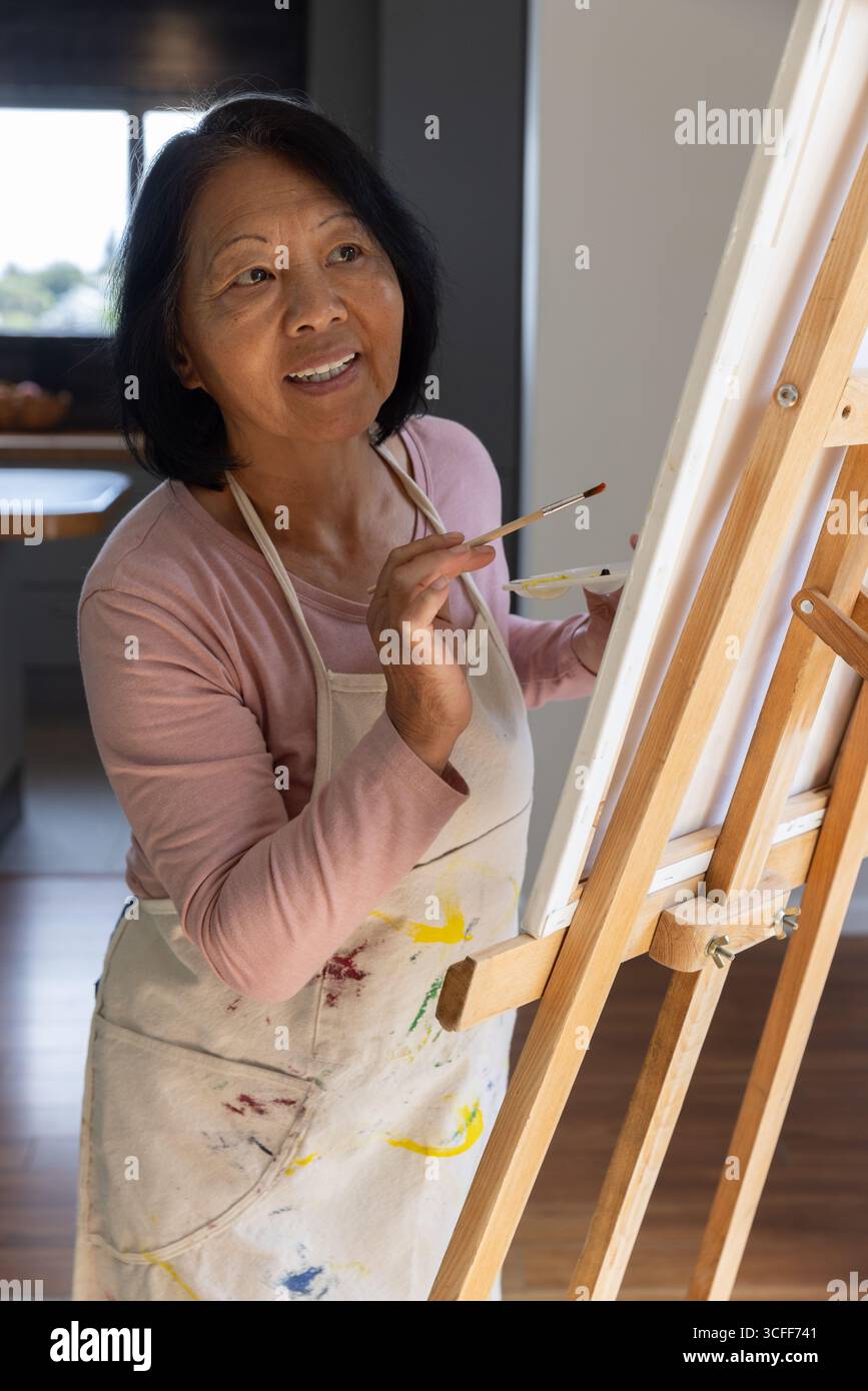 Ältere asiatische Frau malte auf Leinwand im Küchenbereich in lackierter Schürze mit Pinsel Stockfoto
