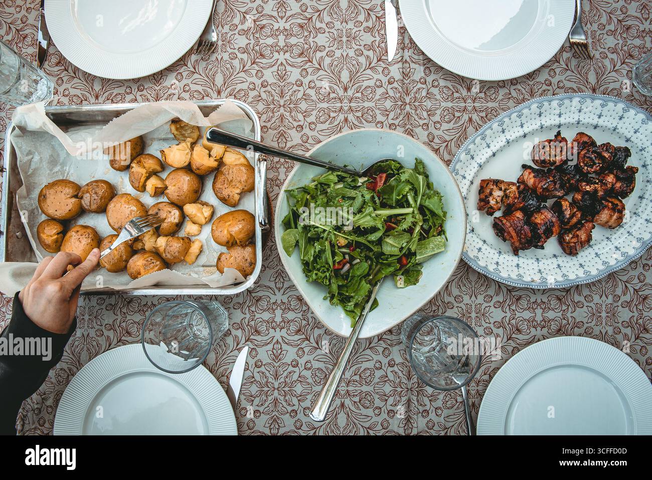Blick von oben auf einen Esstisch mit hausgemachten Gerichten, einschließlich einer Schüssel mit frischem grünem Salat, gebratenen Kartoffeln und gegrillten Schweinebauchspießen. Rustikales f Stockfoto