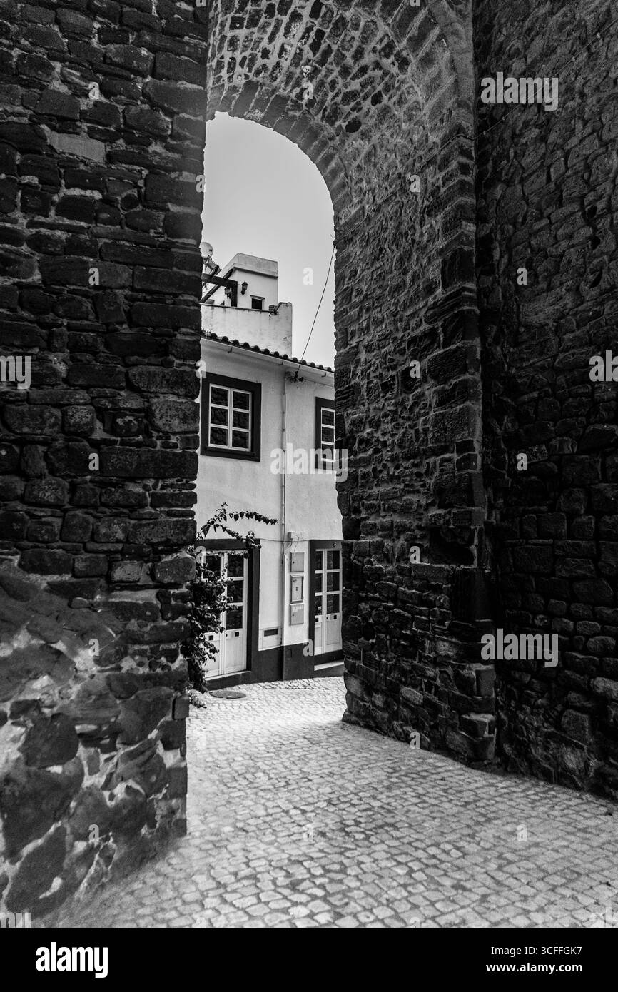 Silves, Algarve, Portugal – 2025.07.04 – Schwarzweiß-Blick durch einen alten Steinbogen, der die kopfsteingepflasterte Straße und die traditionelle weiß getünchte Straße enthüllt Stockfoto