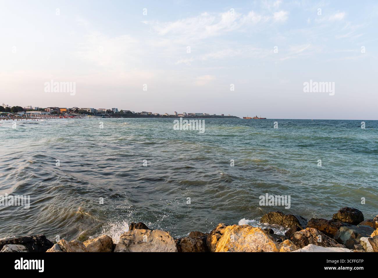 Malerischer Blick auf die Schwarzmeerküste mit Wellen, die Felsen, den Strand und die Skyline der Küstenstadt unter sanftem Abendlicht treffen. Stockfoto
