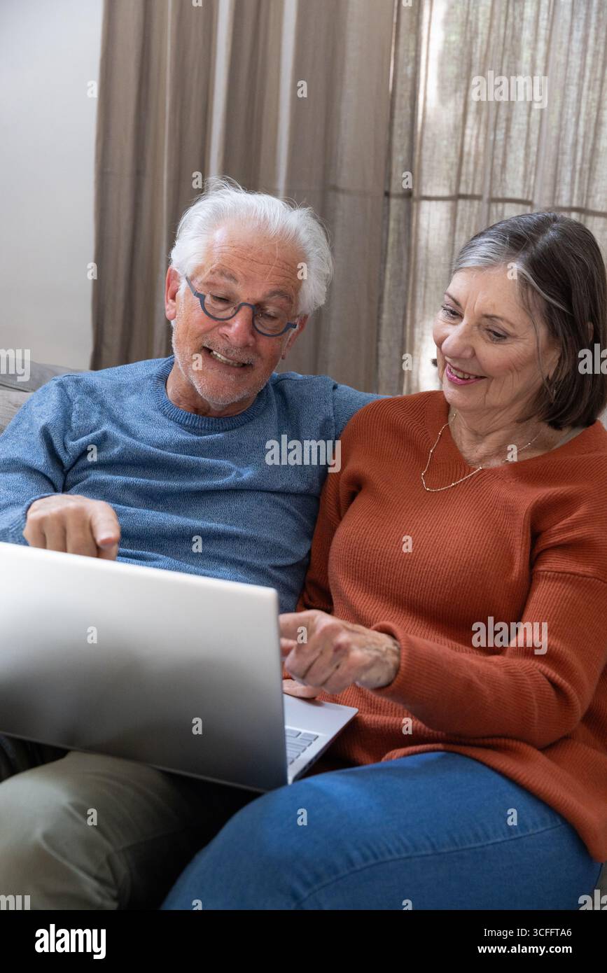 Seniorenpaar lehnt sich nach vorne in Richtung silberner Laptop auf neutralem Sofa im Wohnzimmer zu Hause Stockfoto